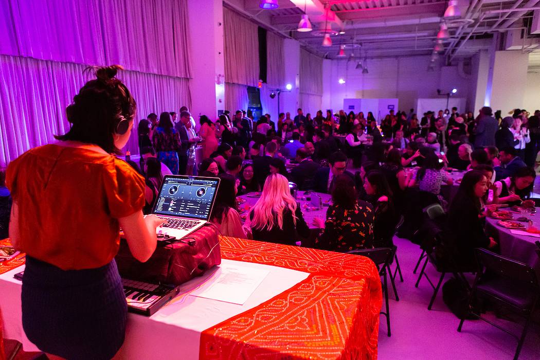 A banquet hall lit with purple and pink lights filled with people at round tables and in the forefront, a DJ on a sound system.