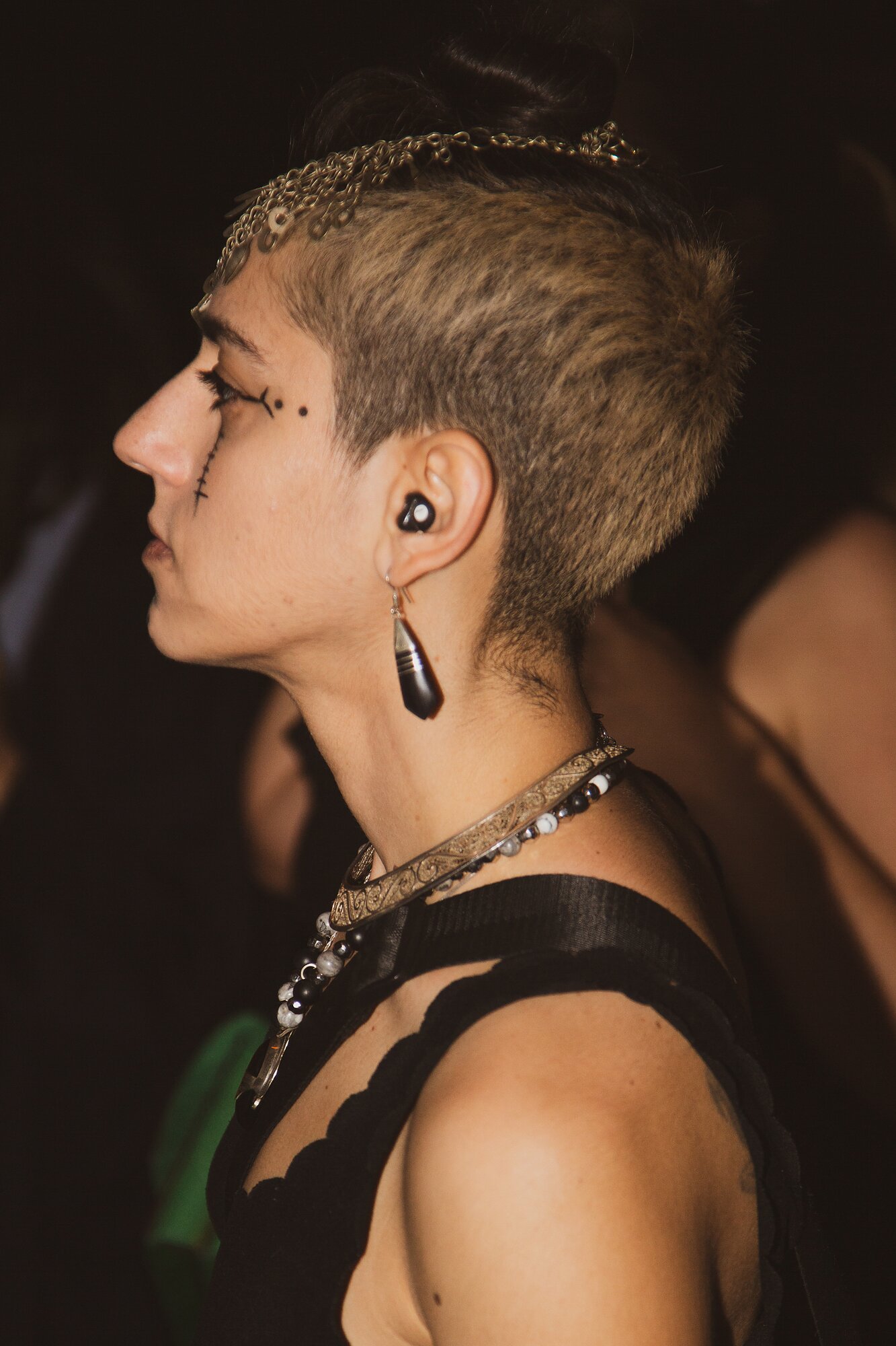 A profile view of a person with a shaved head, elaborate eye makeup, and bold jewelry.