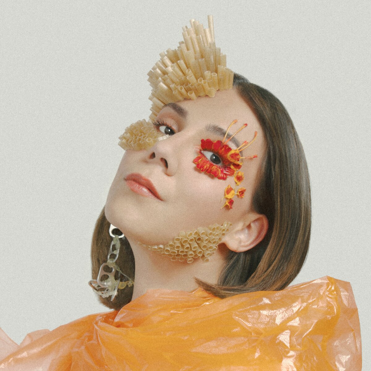 The musician Sirintip seen chest up in three-quarter profile, adorned with flower petals, plastic straws made to look like crystals, can tab earrings, and an orange plastic bag dress.
