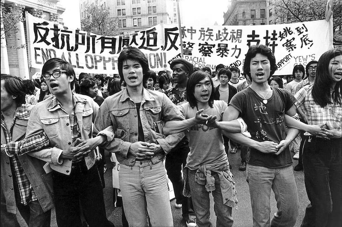 A photograph of a group of young AAPI activists chanting with their arms linked during an anti-police brutality march in NYC in 1975.