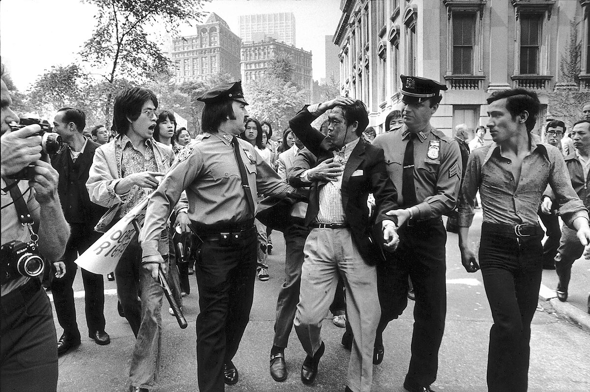 A black and white photograph of police surrounded by AAPI protestors escorting an AAPI man with a bloodied face holding his head.
