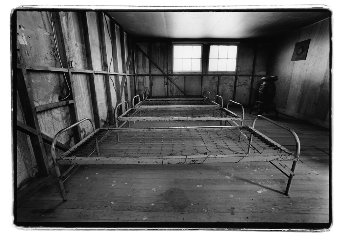 A black and white photo of a row of three empty steel bedframes in a long-abandoned barrack.