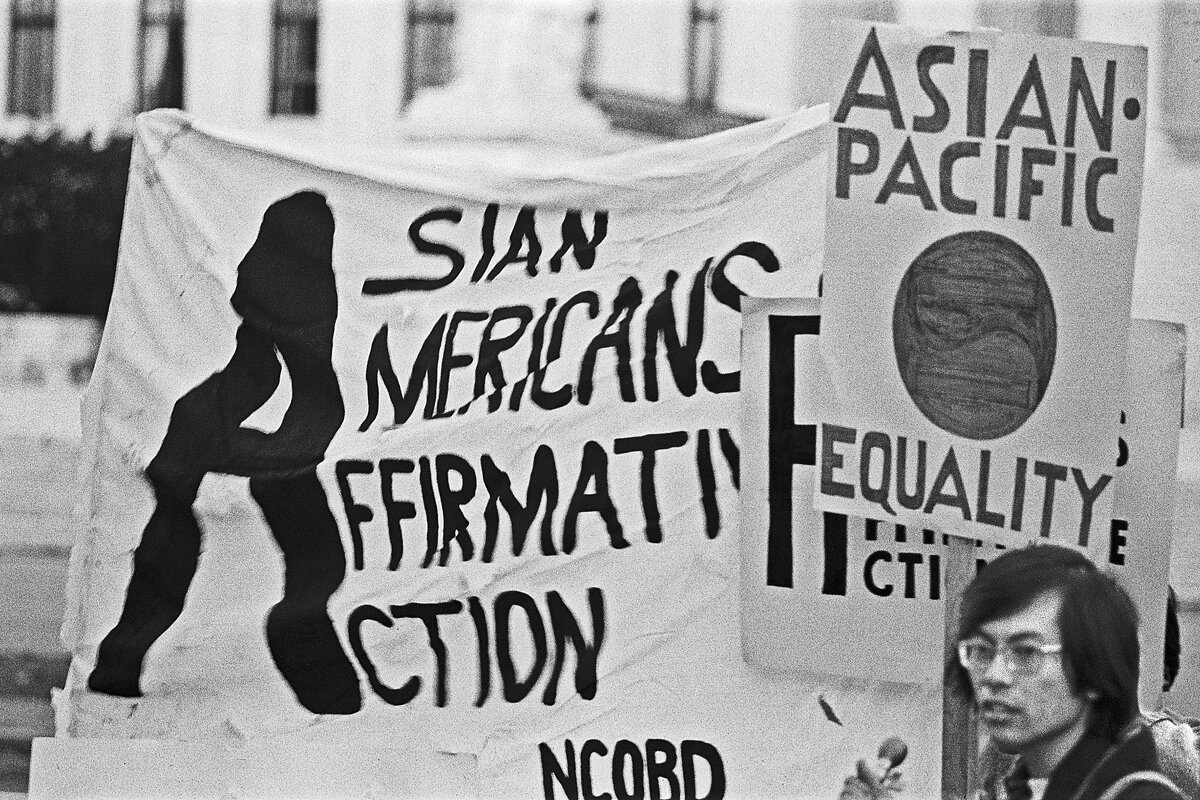 A black and white photograph of Corky Lee in front of protest banners that read "Asian Americans for Affirmative Action" and "Asian-Pacific Equality."