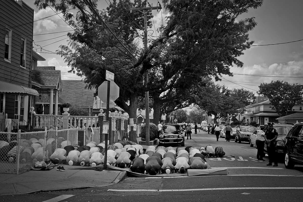 A group of Bangladeshi men praying in the street as an NYPD officer stands watch.
