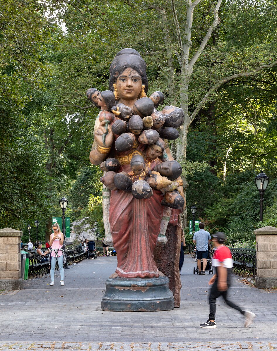 A large bronze statue of an Indian woman holding a flower and dressed in a red sari with dozens of small heads emerging from her body stands in Central Park.