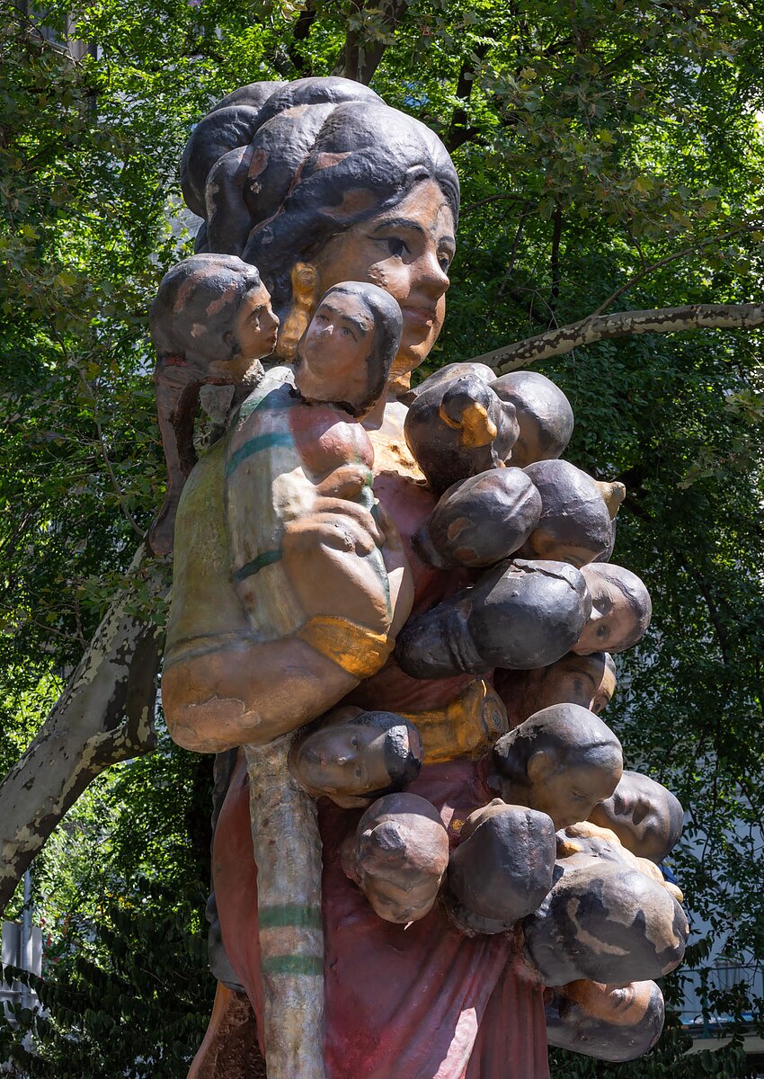 A close-up three-quarter profile view of a large bronze statue of an Indian woman holding a flower and dressed in a red sari with dozens of small heads emerging from her body stands in Central Park.