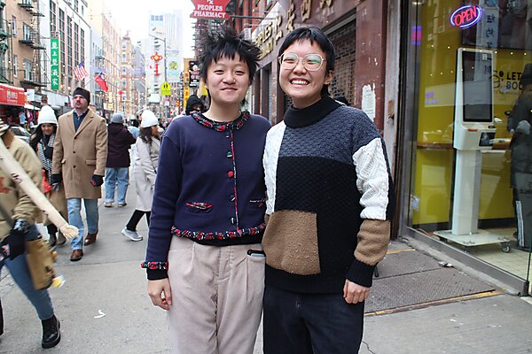 Two Asian people posing in a busy city street. One is wearing glasses and a sweater. The other one has short hair and a cardigan.