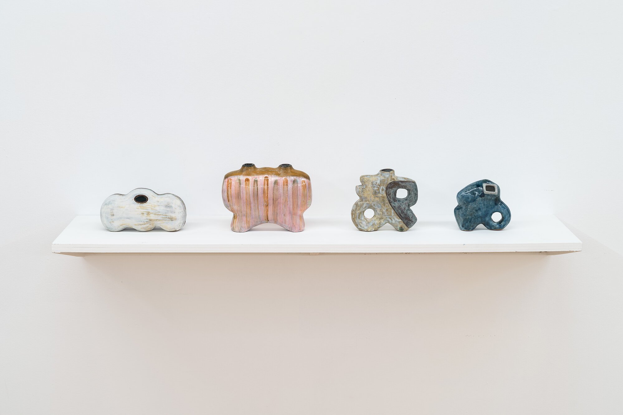 Four ceramic vessels arranged horizontally on a shelf against a white wall.