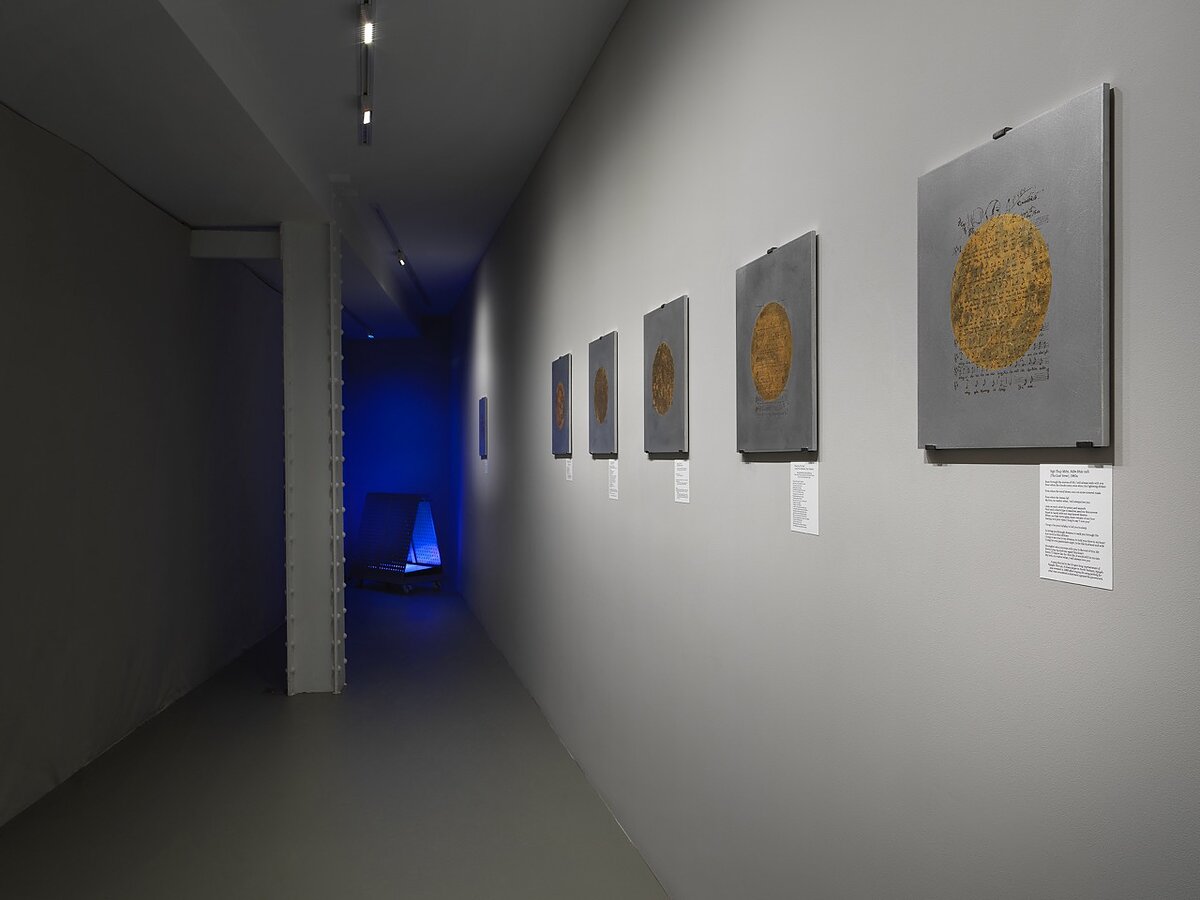 A gallery hallway featuring a series of metallic square works on the walls with gold circles at their centers.