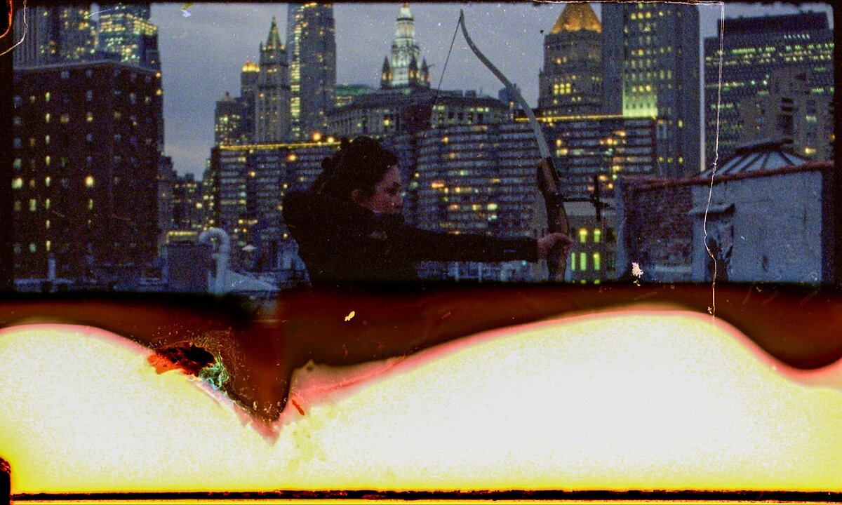A semi-decayed film still of a woman on a city rooftop at sunset shooting an arrow.