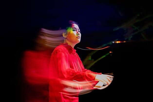 A blurry photograph of a woman in a bright red shirt playing the piano. 