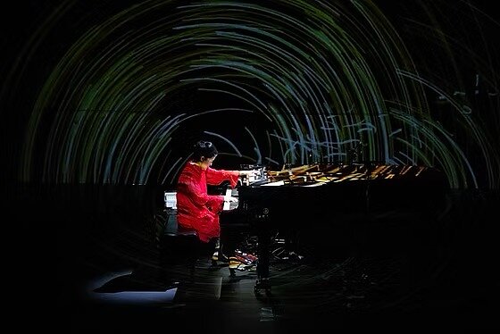 A woman wearing a red dress is seen playing a grand piano on stage with a projection of time-lapsed lights behind her.