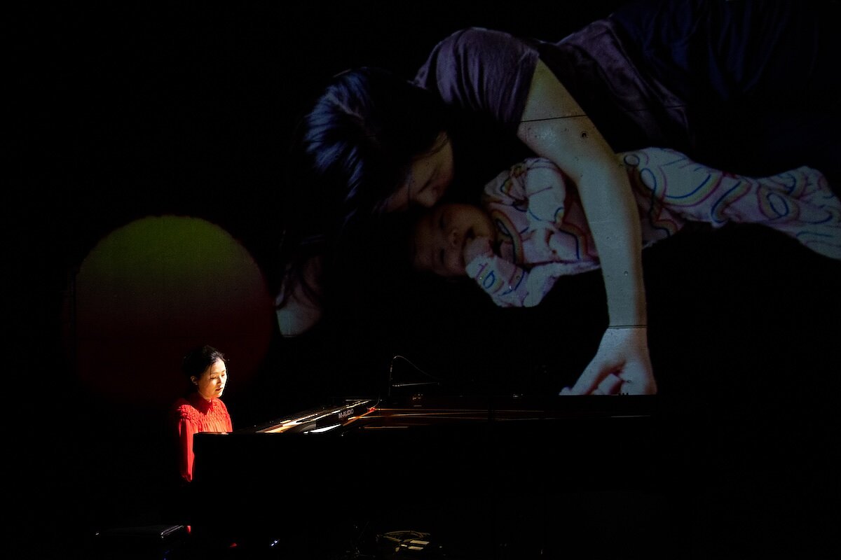 A woman in red sits at a grand piano on a dark stage with a projection of a home video of a mother and baby behind her. 