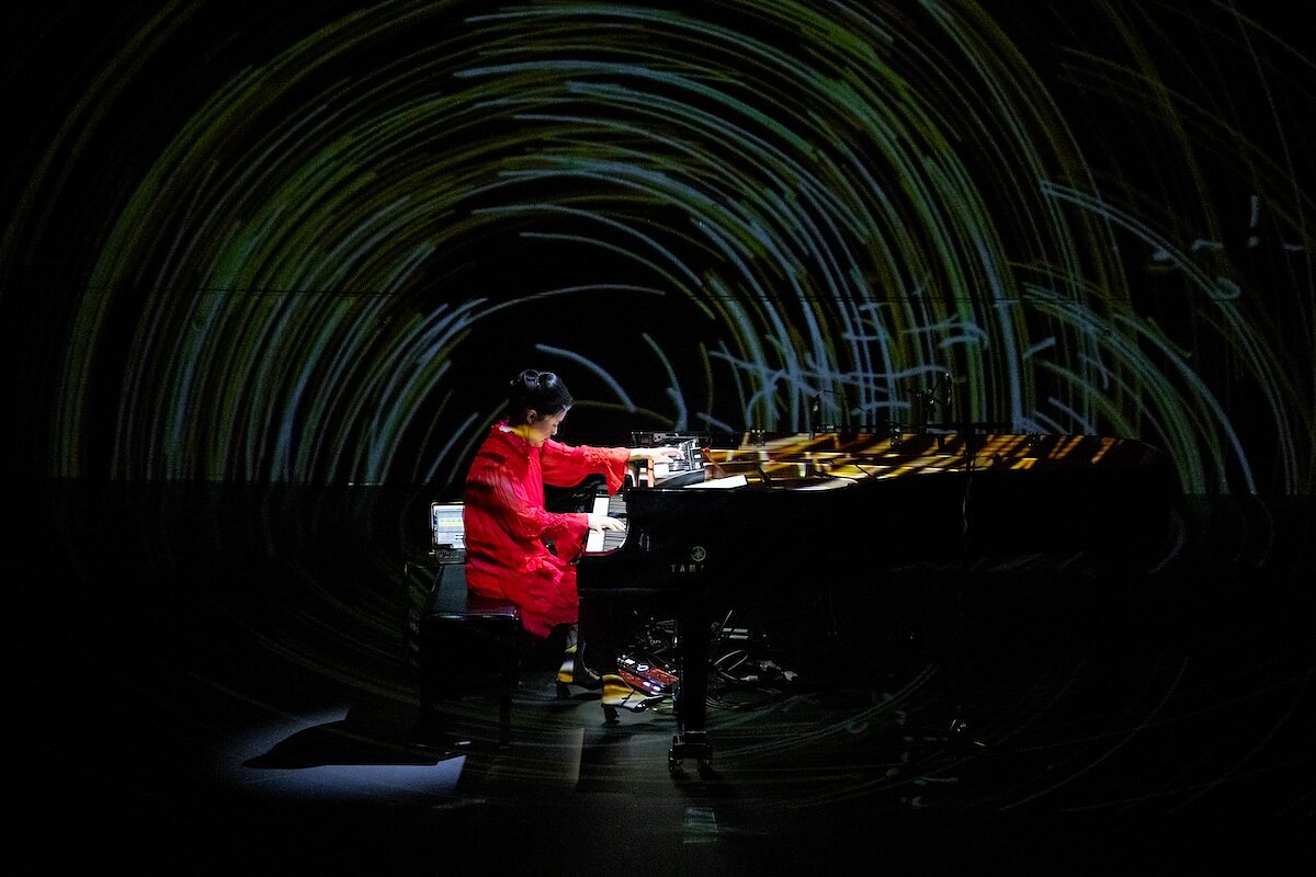 A woman wearing a red dress is seen playing a grand piano on stage with a projection of time-lapsed lights behind her.
