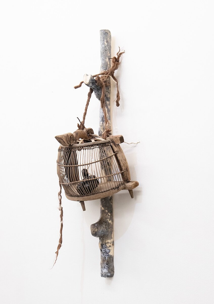 A sculpture made of a wooden stick, birdcage, and rope hangs from a gallery wall. 