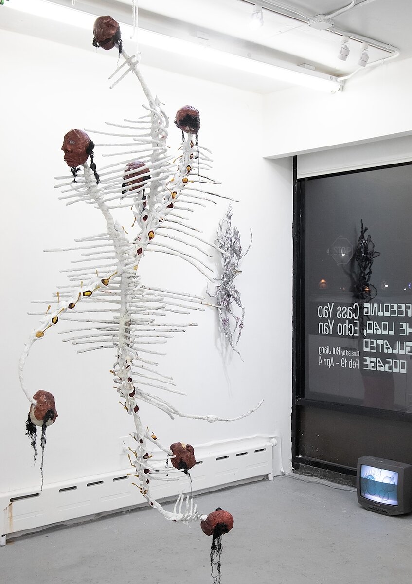 A sculpture resembling a double helix or a spine with clay heads stuck to its ends hangs from a gallery ceiling. Behind it is a spidery silver sculpture hanging from the wall and a television monitor on the floor playing images of microbes. 