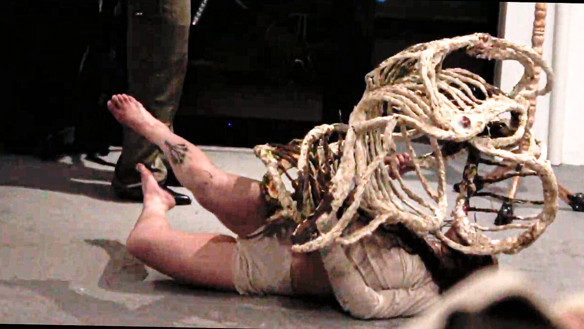 A figure is lying down on a gallery floor, wrestling with a sculpture that resembles a giant ribcage. 