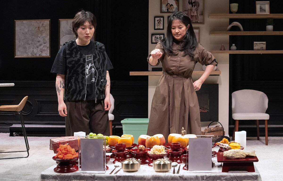 Two woman onstage stand in front of a Jesa table honoring their parents filled with cut fruit.