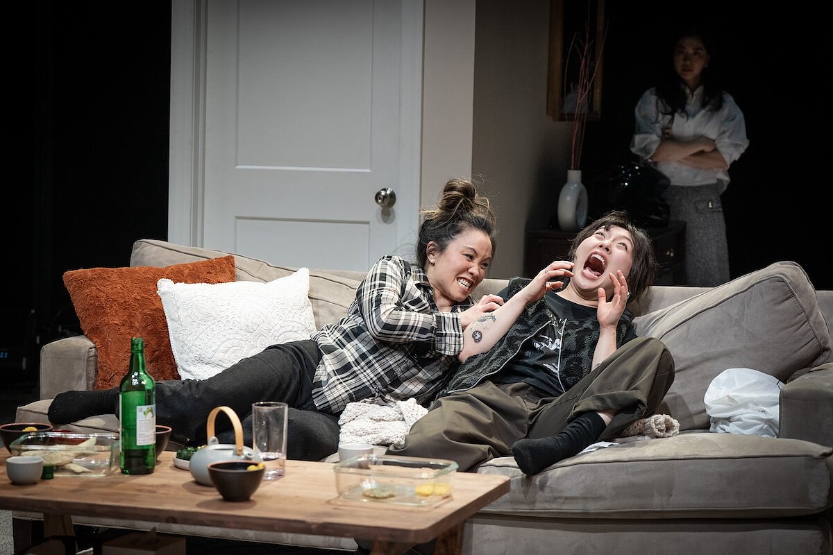 Two women fight onstage on a couch while another woman stands in the shadows behind them with her arms crossed.