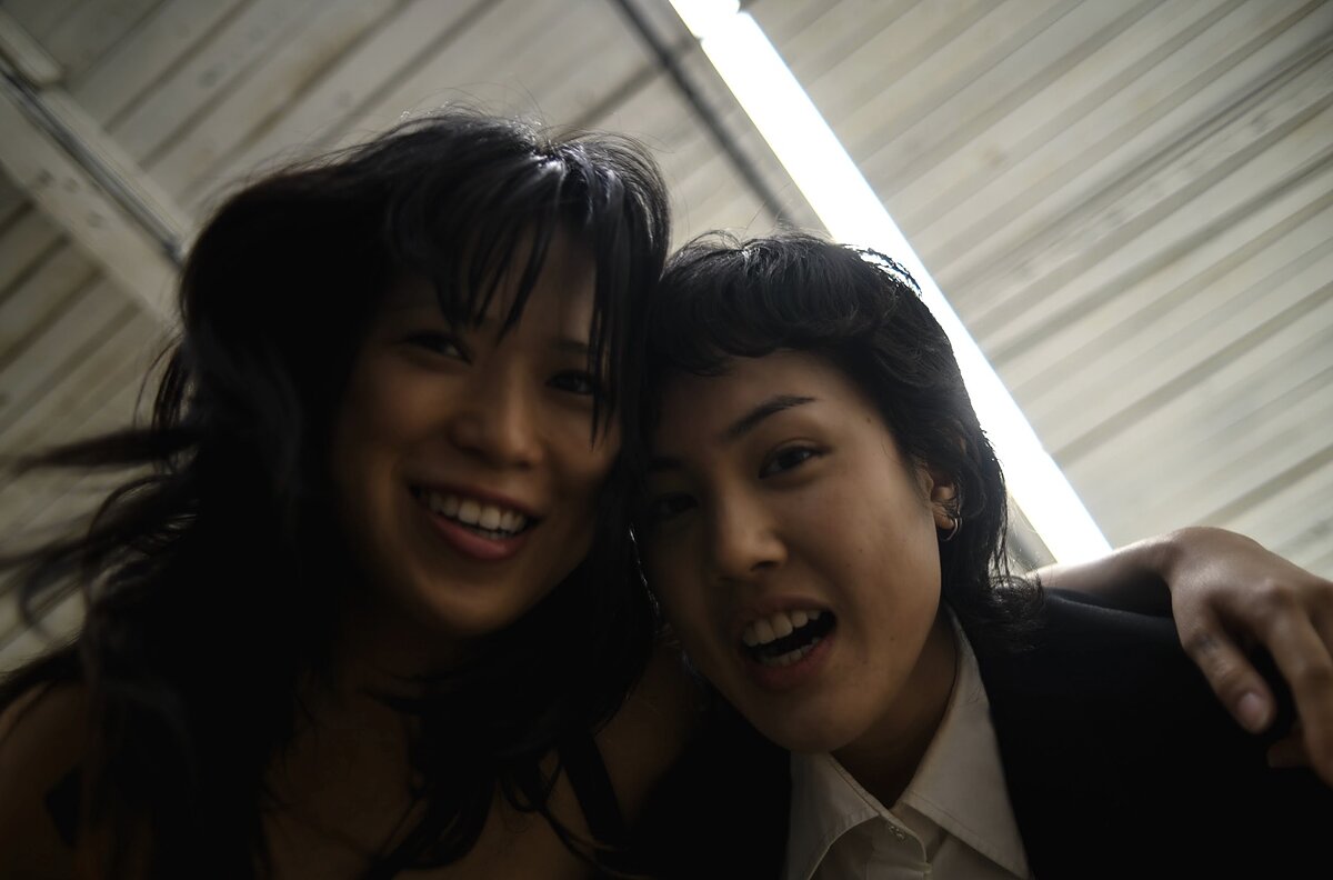Two east Asian women standing closely with their arms around each other, laughing and looking down at the camera. 