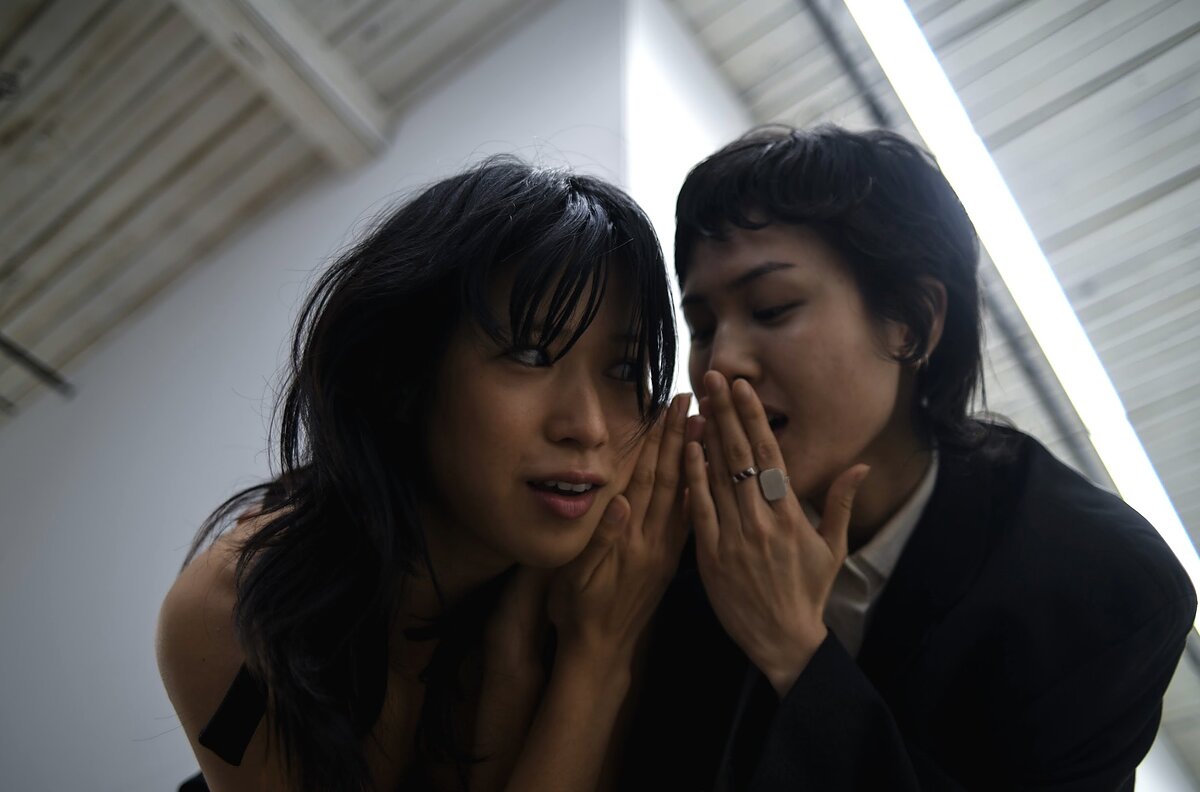 Two east Asian women stand close together, whispering secrets. 