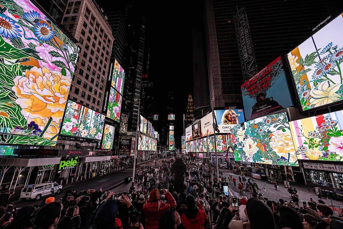 A crowd of people gather around in a city square at night and look up at images on an LED screen depicting colorful animations of florals and patterns.