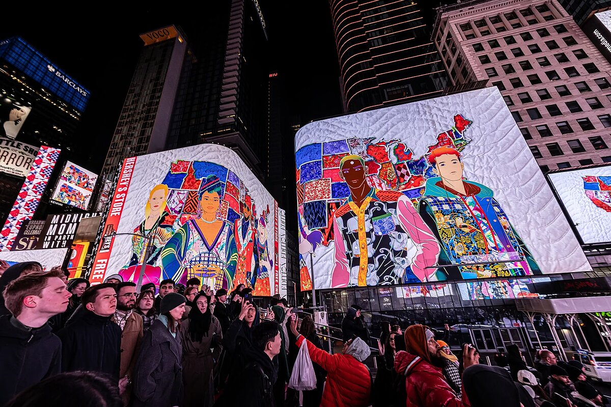 A crowd of people gather around in a city square at night and look up at images on an LED screen depicting colorful animations of people and a map of the United States. 
