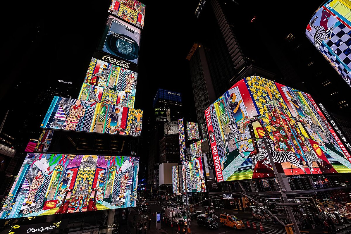 A busy city square at night with LED screens showing a colorful animation of figures walking through brightly patterned rooms amid commercial billboards.