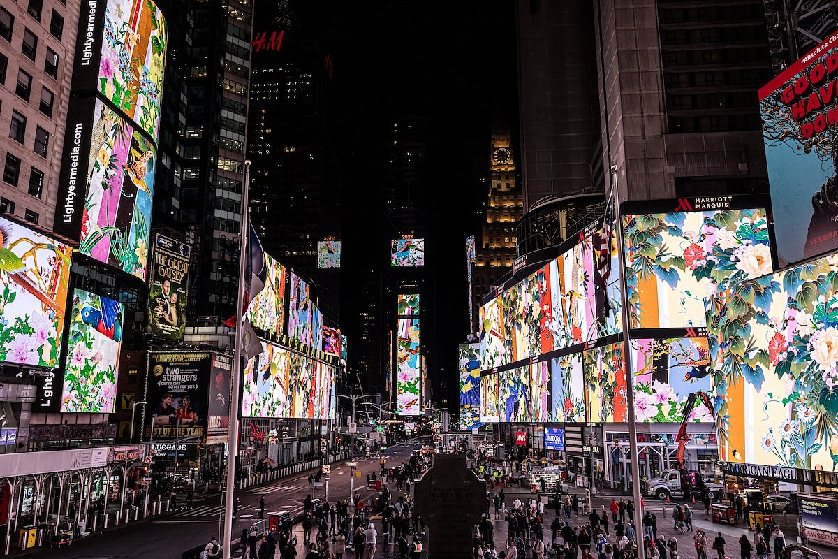 A crowd of people gather around in a city square at night and look up at images on an LED screen depicting colorful animations of florals and patterns.