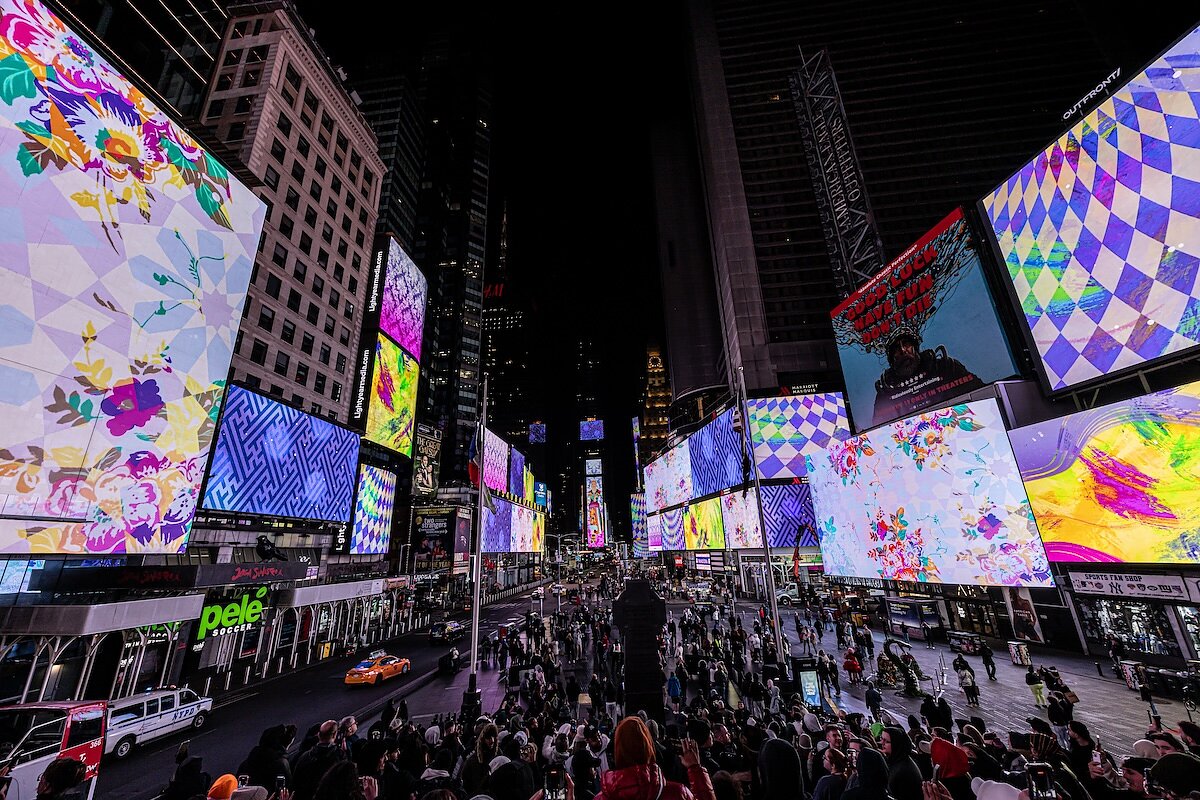 A crowd of people gather around in a city square at night and look up at images on an LED screen depicting colorful animations of florals and patterns.