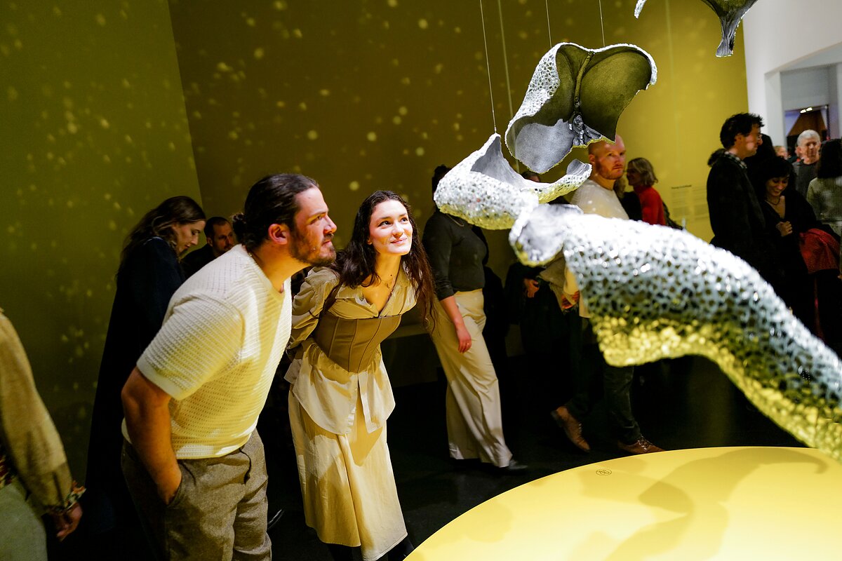 Visitors in a gallery look closely at a mirror-encrusted sculpture that hangs from the ceiling, scattering light throughout the room like a disco ball. 