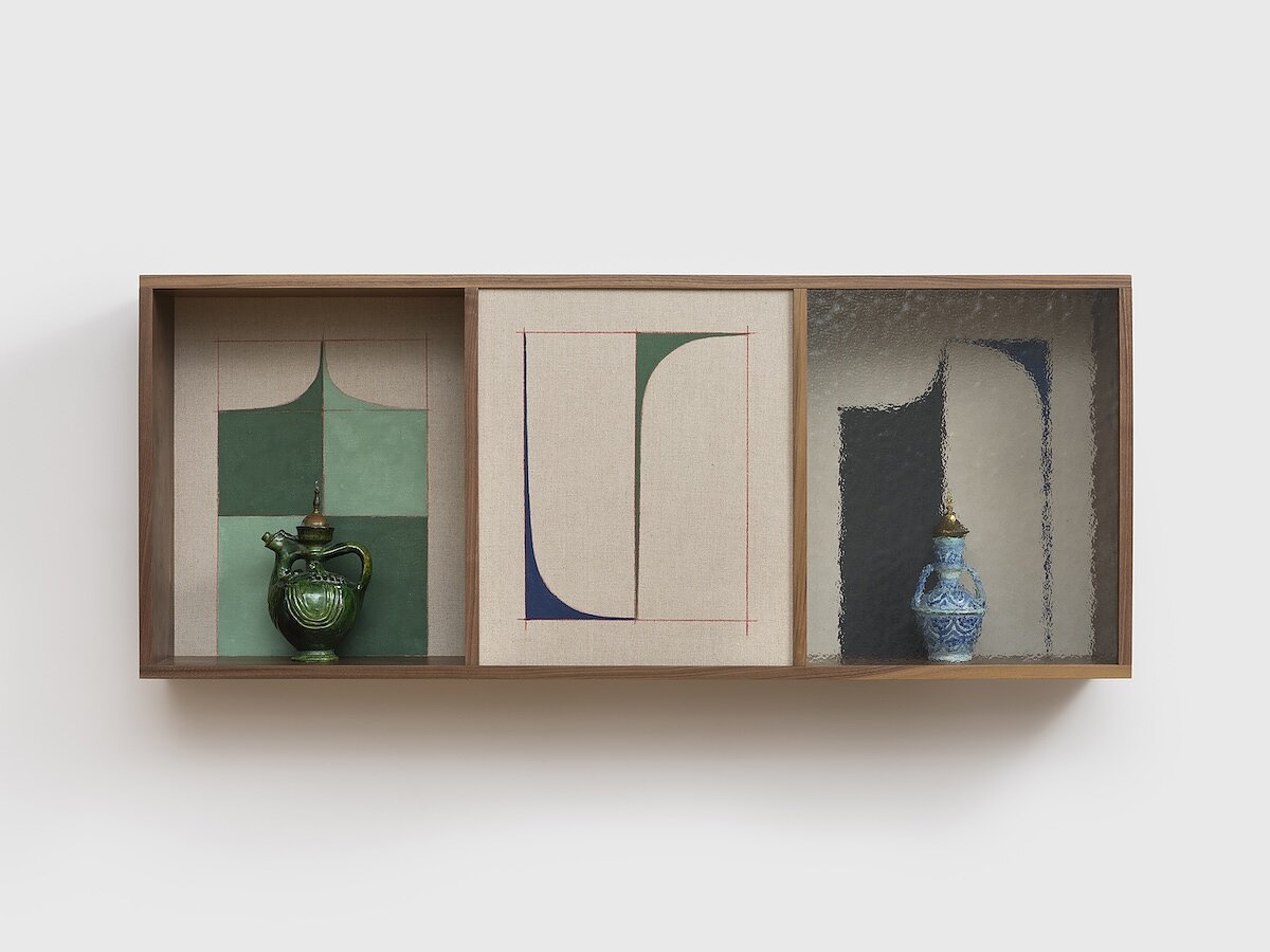 A wooden box hung on a gallery wall displays three canvas painted with blue and green geometric designs. Two ornamental vases are placed within the box, in front of the left and right-most canvases. 