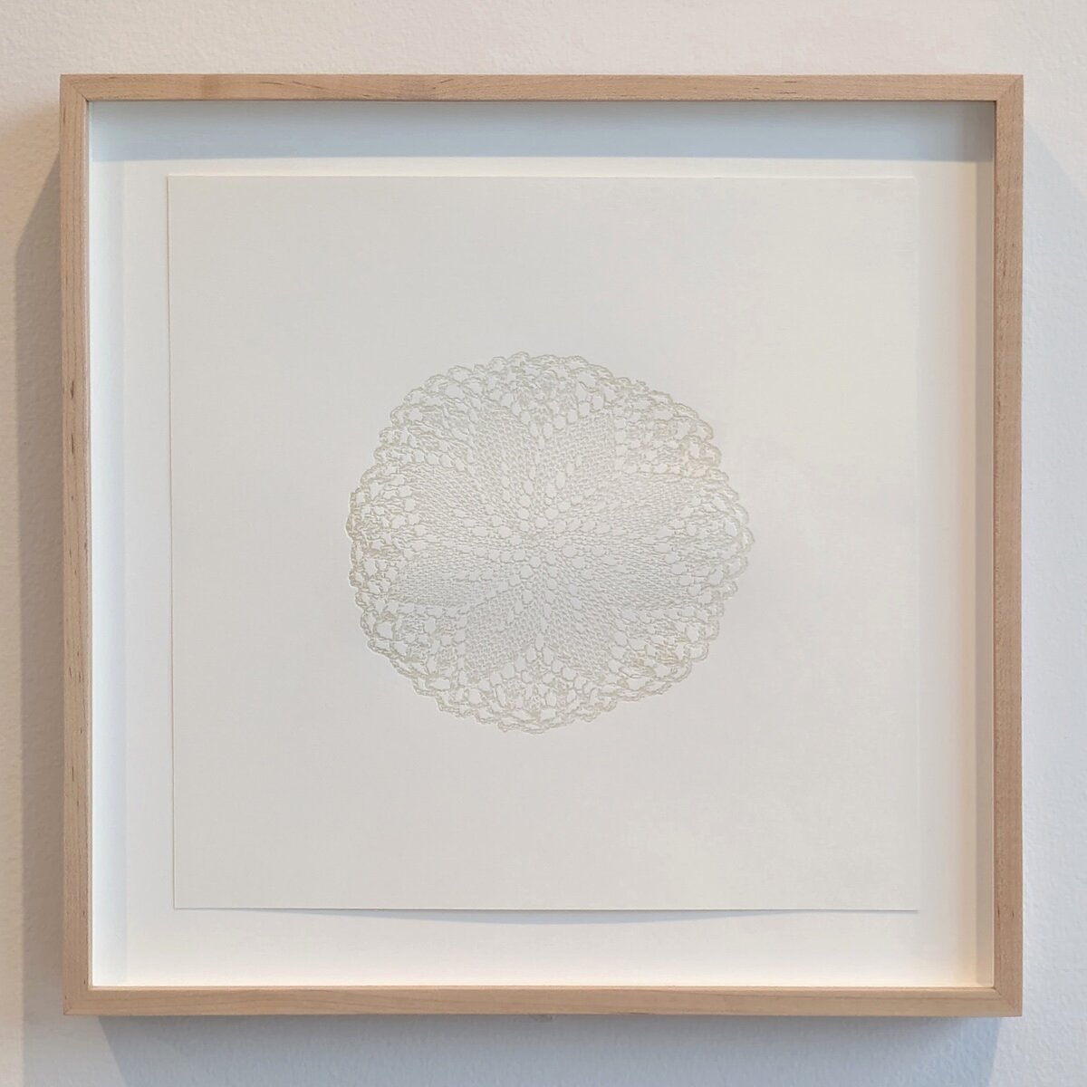 An embossed print of a doily hangs in a square wooden frame. 