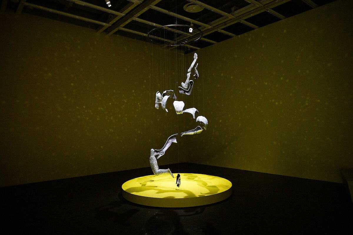 A sculpture covered in mirrored glass hands from the ceiling, casting light onto the yellow gallery walls like a disco ball. 