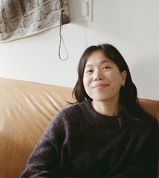 Asian American woman with long hair and gray long sleeved sweater sitting on a leather couch