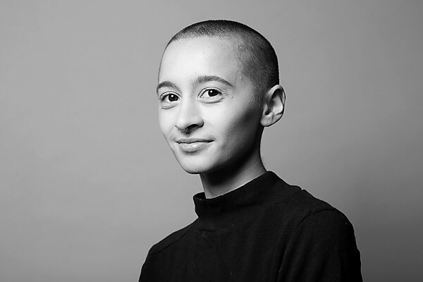 A black-and-white photo of a gender nonconforming person with shaved head and black turtleneck smiling