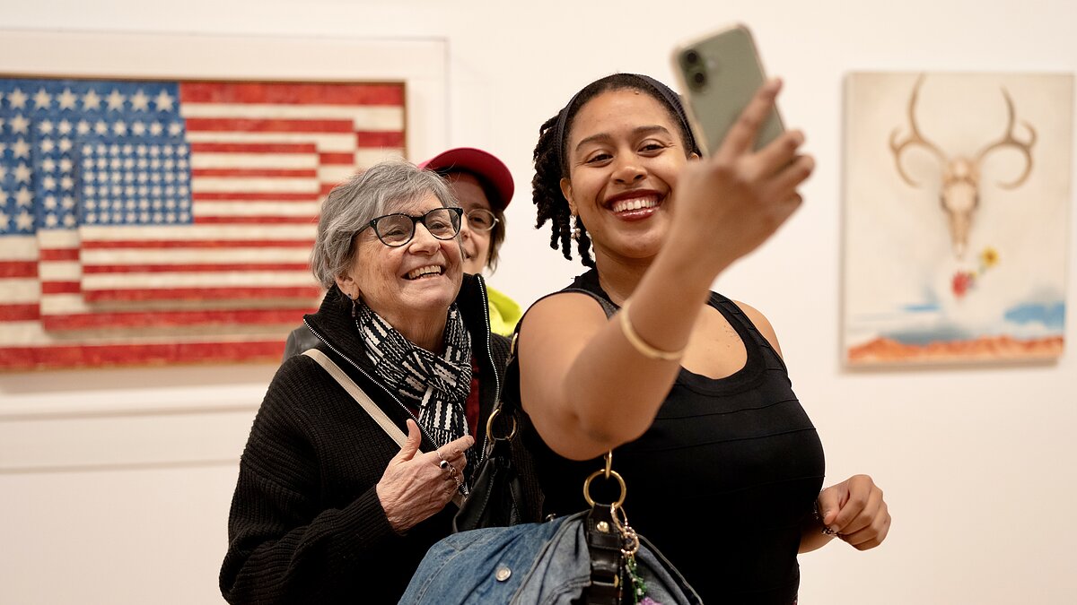 Three museum goers take a selfie in a gallery in front of a painting that features multiple layers of American flags. 