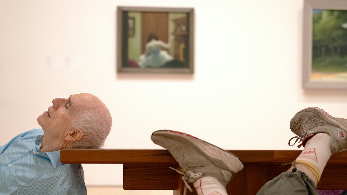 An elderly man rests his head awkwardly on a museum bench in a gallery filled with paintings.