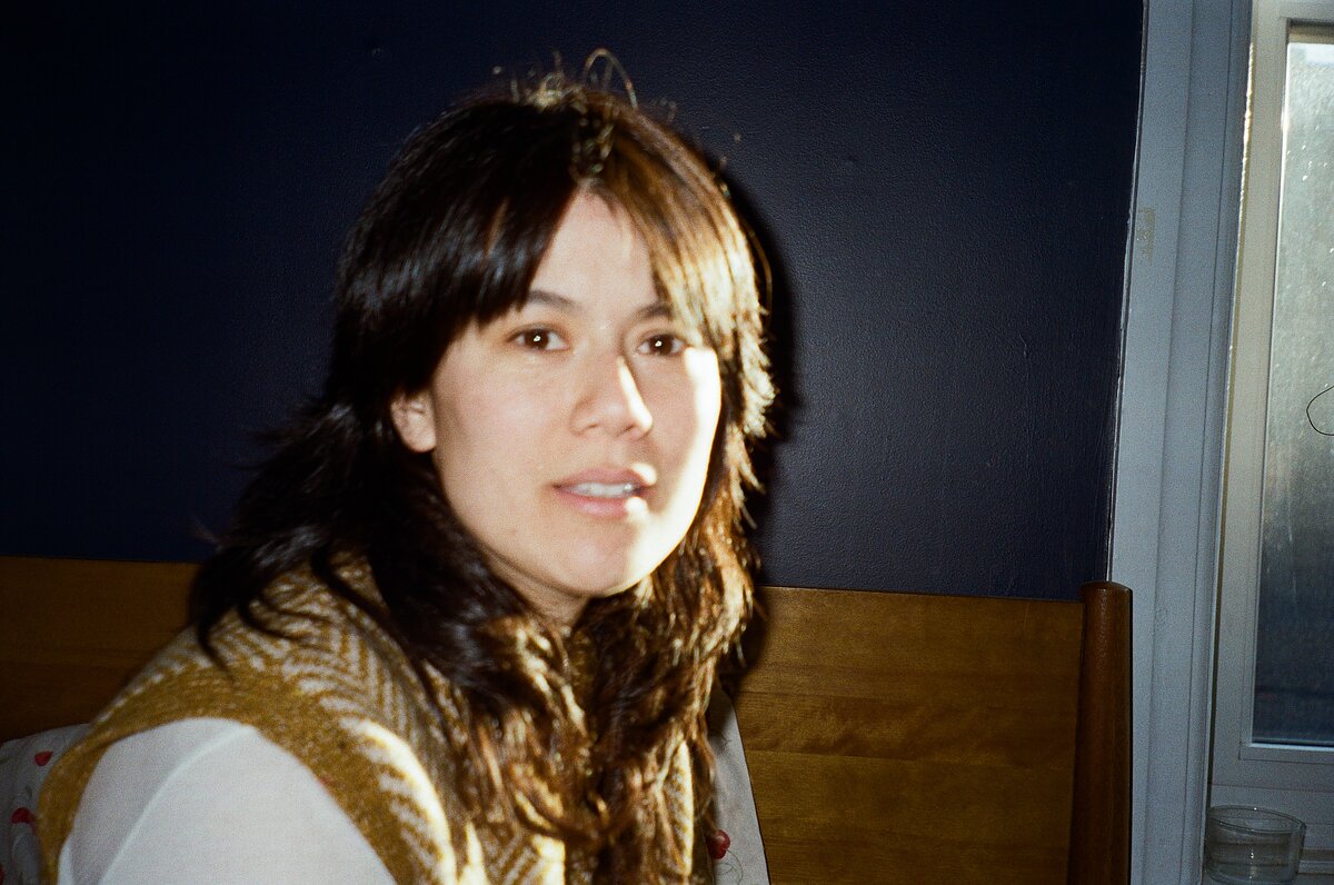Photograph of a woman with choppy bangs wearing a yellow and white knit sweater vest looking at the camera. 