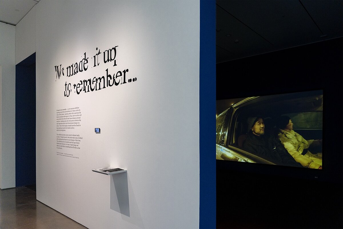 A white gallery wall with text that reads "We made it up to remember..." in a glitchy font. An iPod playing a video is hung up on the wall, behind which we see a dark blue gallery screening a film featuring two figures in a car. 