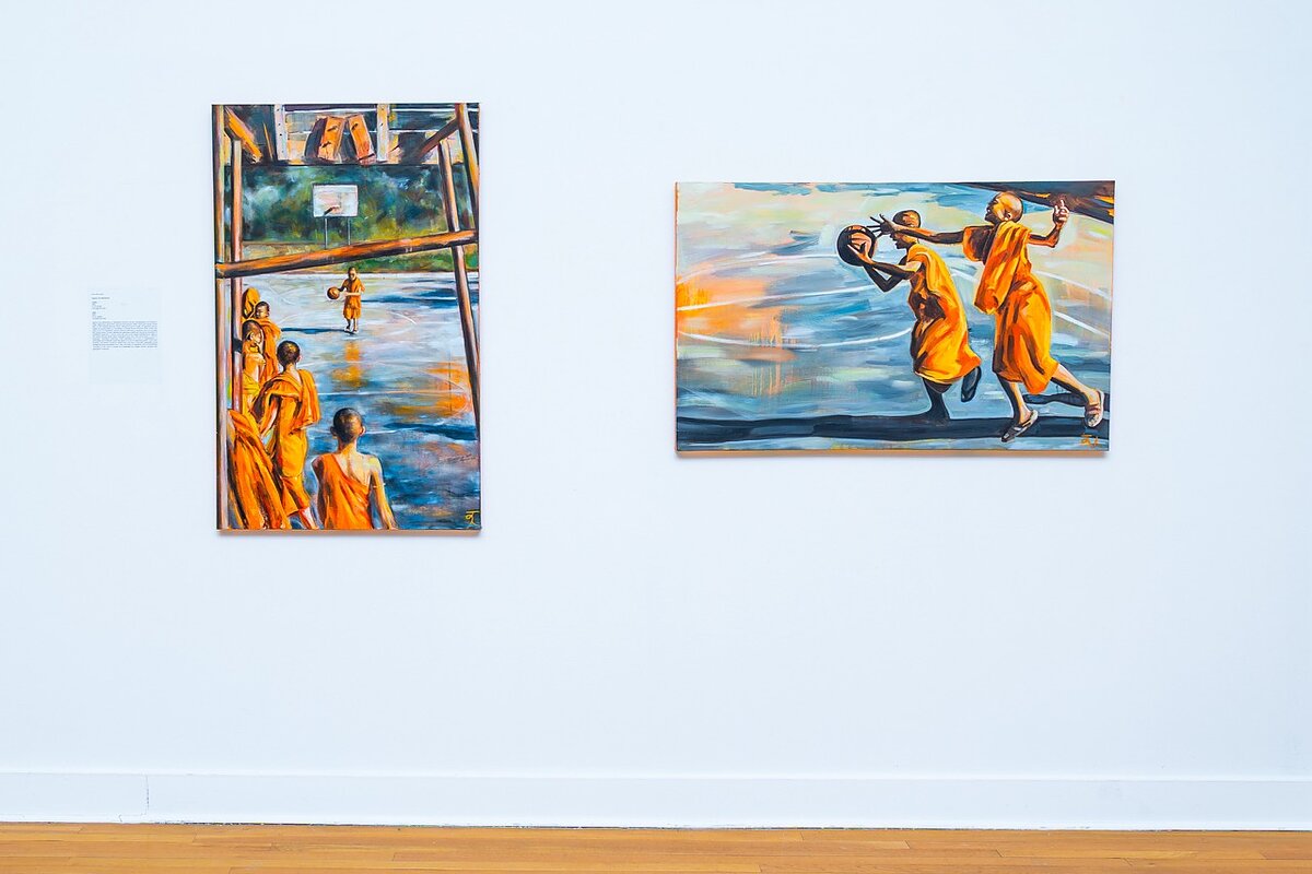 Two paintings hang on a gallery wall featuring monks in orange robes playing basketball. 