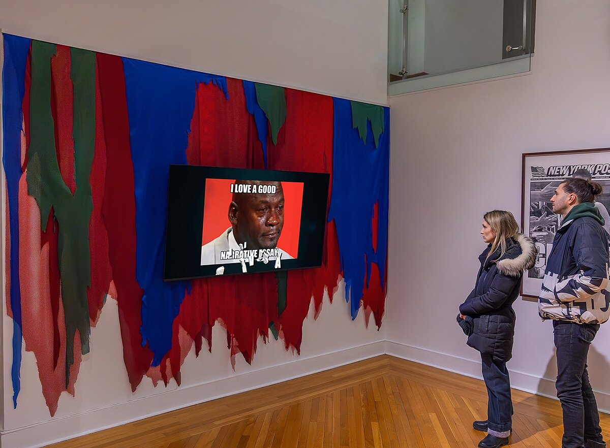 A television screen hung on a colorful gallery wall shows a video art work featuring a meme of the basketball player Michael Jordan. 