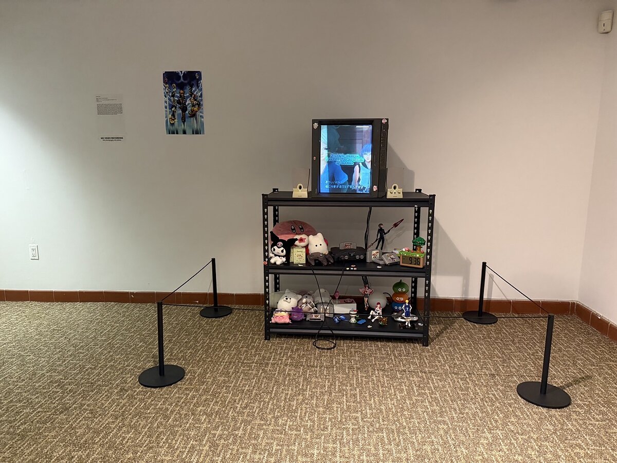 A sculpture featuring a sideways television monitor on a display shelf with plushies and video game ephemera in a gallery. 