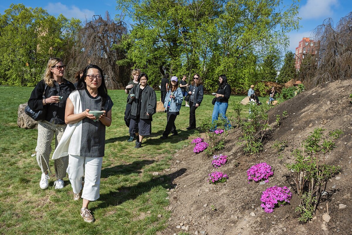 A woman holding a small bowl leads a line of people around a giant mound of dirt with purple flowers. 