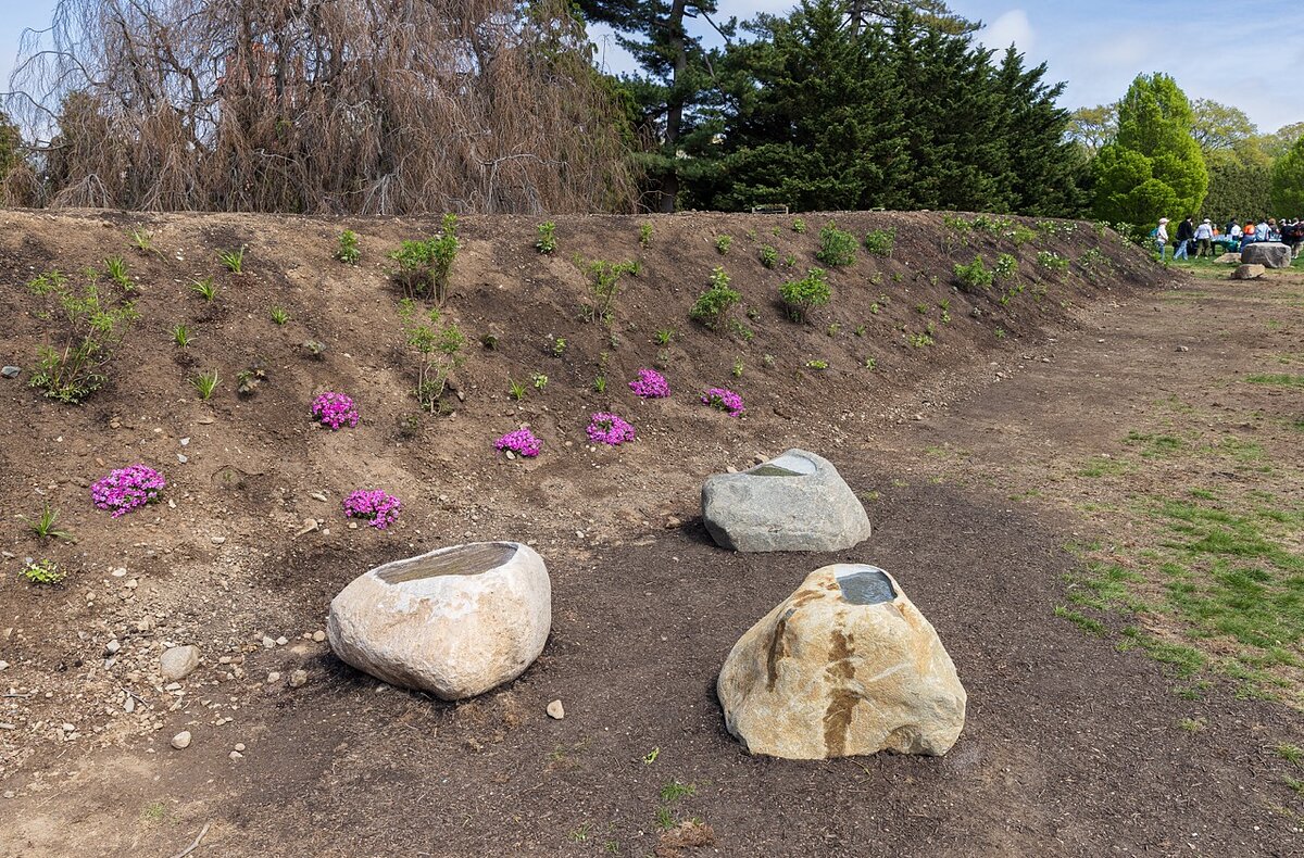 Three stones with shallow basins carved into their tops sit next to a giant mound of dirt with purple flowers. 
