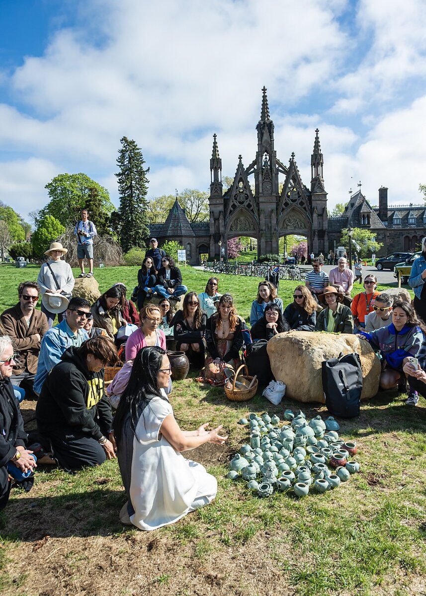 A circle of people sitting on a grassy lawn encircle a pile of small celadon ceramics. Behind them is a cemetery gate. 