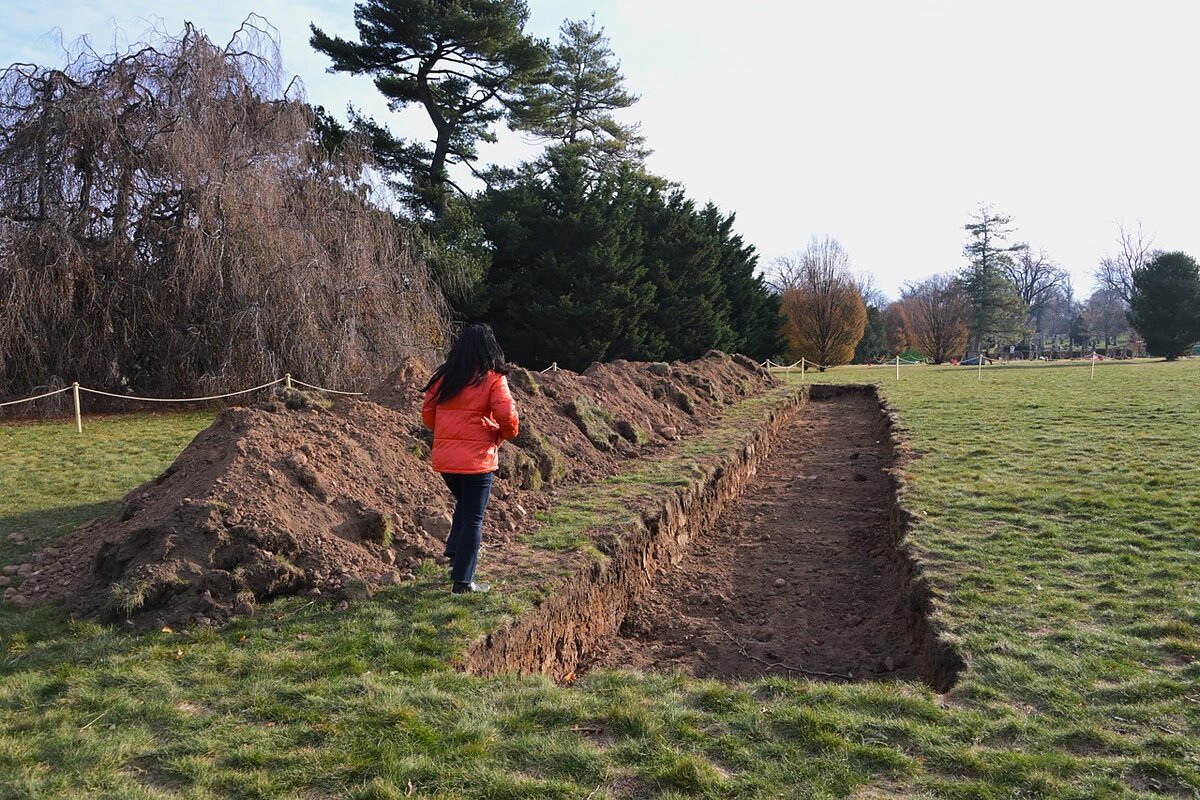 A woman wearing a red puffer jacket stands between a giant rectangular ditch and its corresponding pile of dirt on a grassy lawn. 