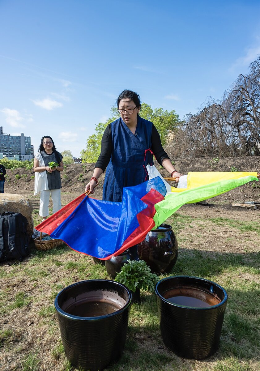 A woman standing outside holding a blue, red, green, and yellow fabric banner over large ceramic vessels. 