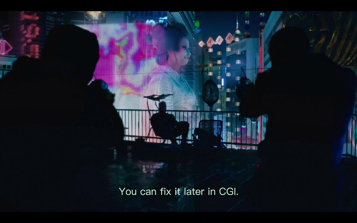 A screenshot from the film "Bladerunner" featuring two assassins on a futuristic rooftop aiming their guns at a seated figure looking at a giant hologram of a geisha.