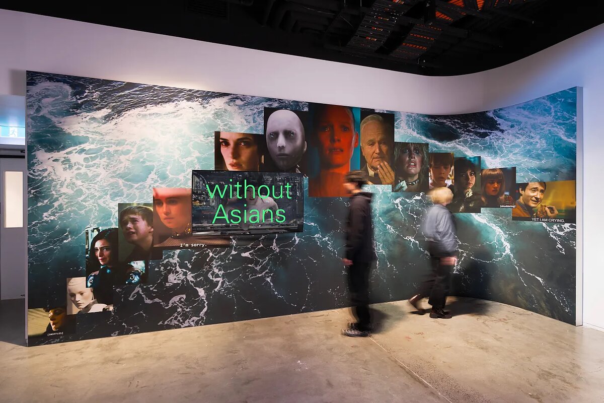 Two blurry people in a gallery whose walls feature ocean waves and screenshots of faces from a variety sci-fi films and one with the words "without Asians" in bright green font.